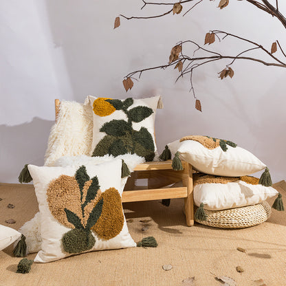 A styled arrangement of decorative throw pillows with textured botanical tufted designs in cream, olive green, and mustard yellow, displayed on a small wooden bench and floor against a neutral wall with dried branches overhead.