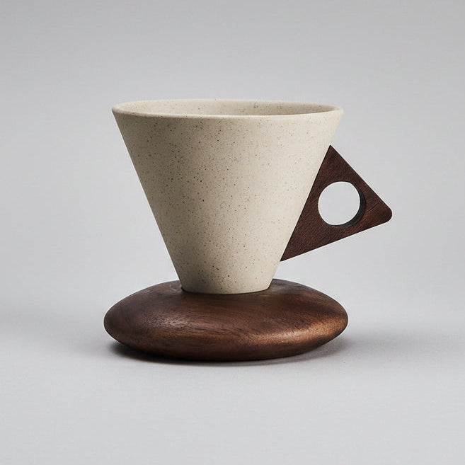 A minimalist ceramic pour-over coffee dripper with a speckled off-white cone body, a dark brown triangular handle with a circular cutout, and a matching dark brown wooden base, set against a seamless light gray background.