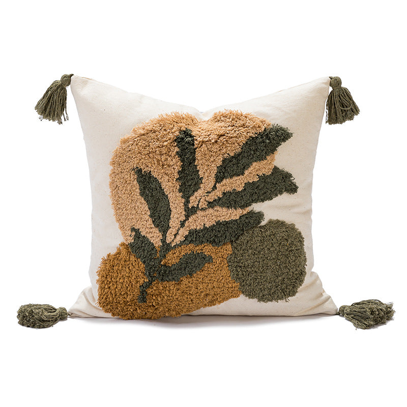 A square off-white throw pillow featuring a raised, shaggy botanical motif in tan, olive green, and dark forest green, finished with four dark green tassels.