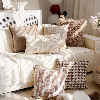 A styled living room scene with multiple decorative throw pillows arranged on a cream sofa and floor. The pillows include textured cream, taupe, and brown patterns, including a fluffy pillow, a geometric pillow, and a houndstooth-style pillow.
