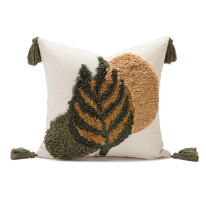 A square throw pillow with a cream-colored base featuring a raised, textured abstract design of a leaf in olive green and tan, with matching olive green tassels on the corners, set against a plain white background.