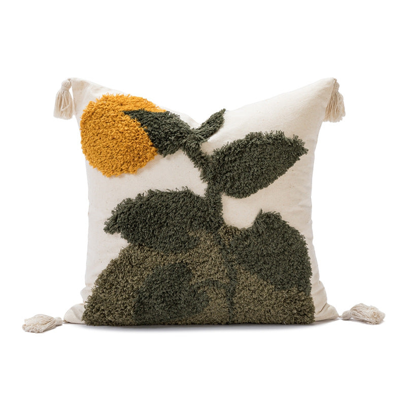 A square throw pillow with a textured, shaggy surface featuring a stylized floral design in dark green and mustard yellow on an off-white background, finished with cream tassels at the corners.
