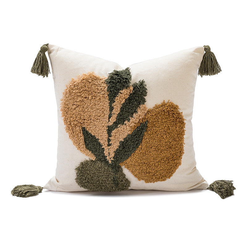 A square throw pillow with a cream-colored base featuring a raised, shaggy abstract floral design in tan and dark olive green. The pillow is finished with tassels on all four corners, photographed against a seamless white background.